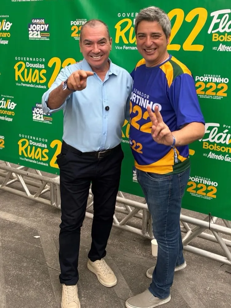 Paulo Bastos e outro candidato posando sorridentes em frente a painel de campanha da direita fluminense